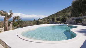 A curving infinity pool with clear blue water, surrounded by a white tiled deck, set against a backdrop of a mountainous landscape and a clear sky. - Hillside villa with pool for 5 guests in Monopoli (Monopoli)