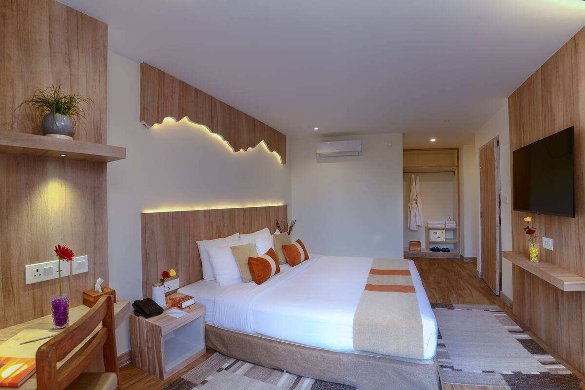 A modern hotel room with a large bed, wooden paneling, a flat-screen TV, a bedside table with a lamp, and a small desk with a chair.