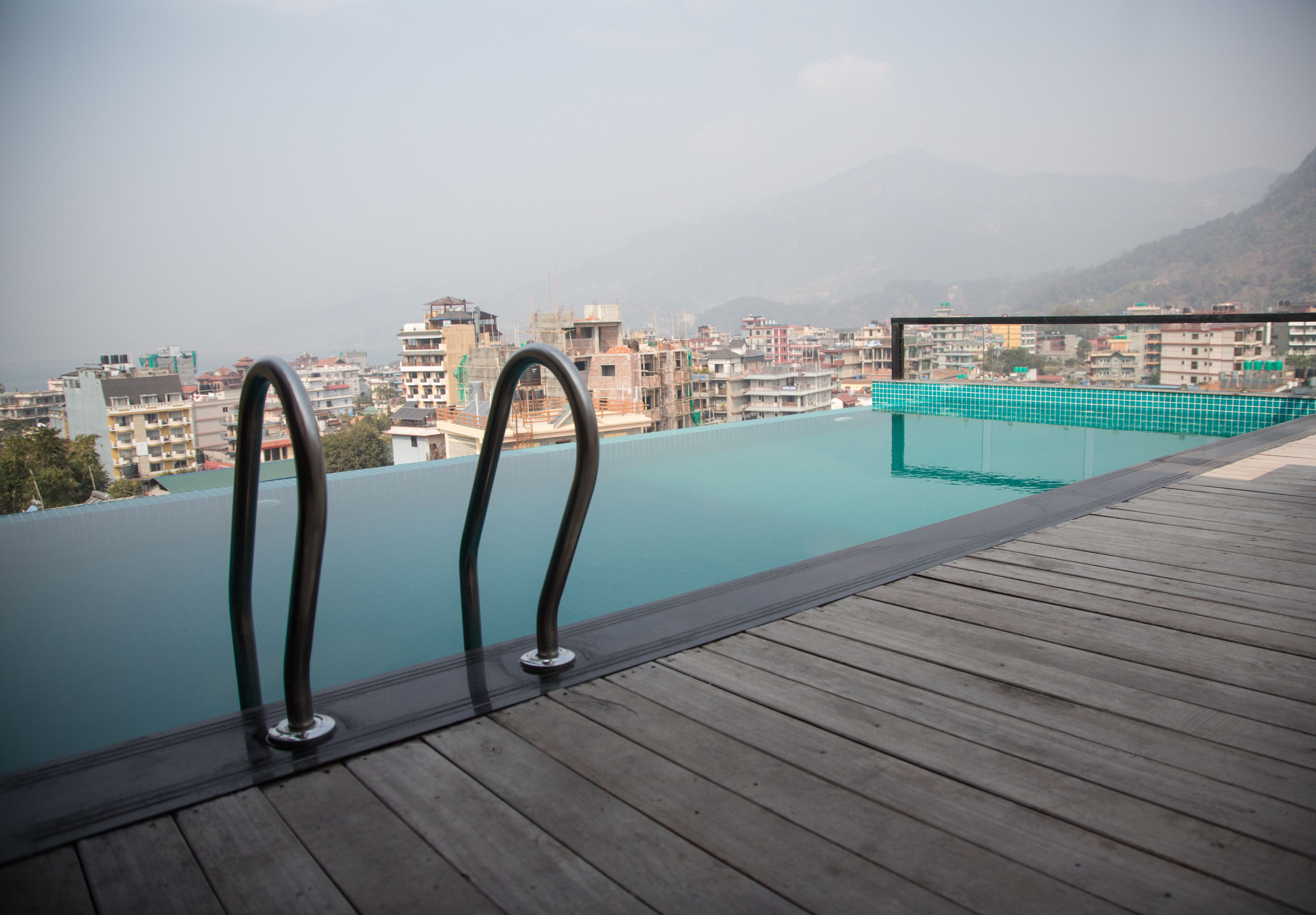 Una piscina infinita con barandillas de metal, una terraza de madera y un paisaje urbano con montañas al fondo.