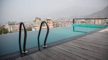 Una piscina infinita con barandillas de metal, una terraza de madera y un paisaje urbano con montañas al fondo.