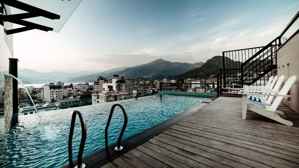 A rooftop infinity pool with a wooden deck and white lounge chairs, overlooking a mountainous town. - Aabas Pokhara by Barahi Hospitality (Pokhara)