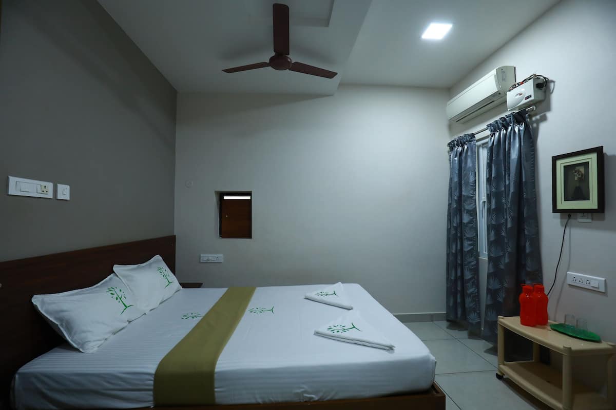 Een net opgemaakt bed met witte lakens en groene accenten, een plafondventilator, een airconditioning en een klein nachtkastje met rode flessen, plus een lijst met een foto aan de muur.