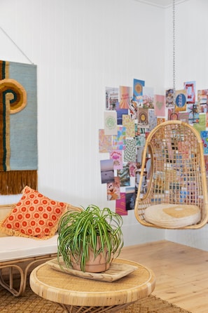 Una habitación con una silla de mimbre, una mesita con una planta en maceta, un sofá con cojines estampados y una pared decorada con un collage de fotos.