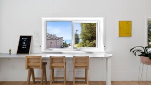 A room with a white desk, two wooden chairs, a window with a view of a building and trees, and a yellow artwork on the wall.