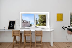 Habitació amb escriptori blanc, dues cadires de fusta, finestra amb vista a un edifici i arbres, i una obra d'art groga a la paret.