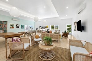 A spacious living area with a dining table, wicker furniture, a round coffee table, and a television mounted on the wall. - The Pink Lodge (Lorne)