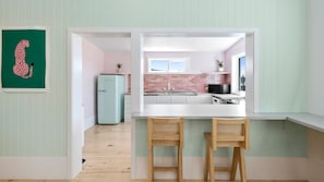 Une cuisine moderne avec un plan de travail blanc, deux tabourets en bois, un réfrigérateur de style vintage et un mur de fond en briques.