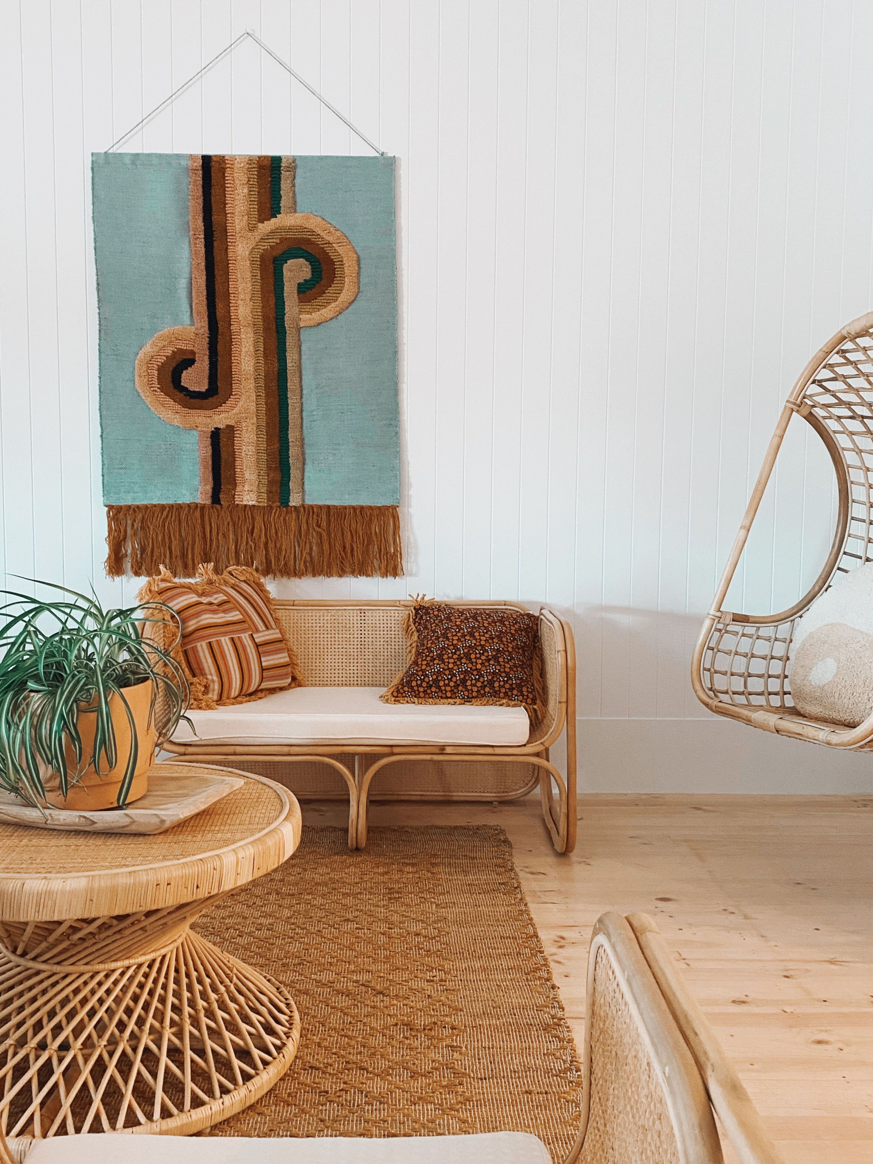 Ein Zimmer mit einem gewebten Rattan-Sofa, einer hängenden Wandweberei, einem Korbstuhl und einem gewebten runden Tisch mit einer Topfpflanze.