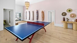A room with a ping pong table, a wooden floor, a pink wall with surfboards, and a ceiling light.