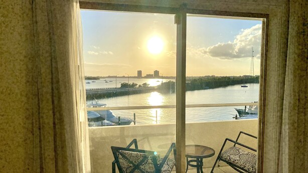 A balcony with a view of a river and boats, a table, and two chairs.