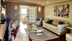 A modern living room with a sofa, coffee table, and chairs. There is a dining area with a table and chairs. The room has a large window and artwork on the walls.