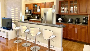 A modern kitchen with wooden cabinets, a stainless steel refrigerator, a microwave, and a coffee maker. There are three bar stools and a television.