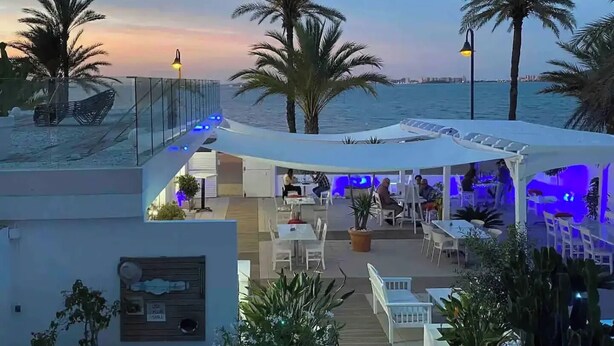 Eine Dachterrasse mit Sitzgelegenheiten im Freien, Palmen und Blick auf das Meer.
