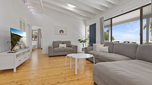 Un salon moderne avec une grande fenêtre, un téléviseur à écran plat, un canapé gris, une table basse blanche et un plancher en bois.