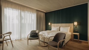 A modern hotel room with a wooden floor, a large bed with white linens, two chairs, a small table, and a nightstand with lamps. - Landhotel Bohrerhof (Hartheim am Rhein)