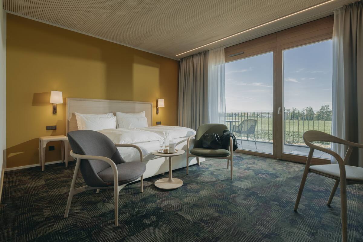 Una habitación de hotel con una cama grande, dos sillas, una mesita y vistas al exterior a través de una puerta corrediza.