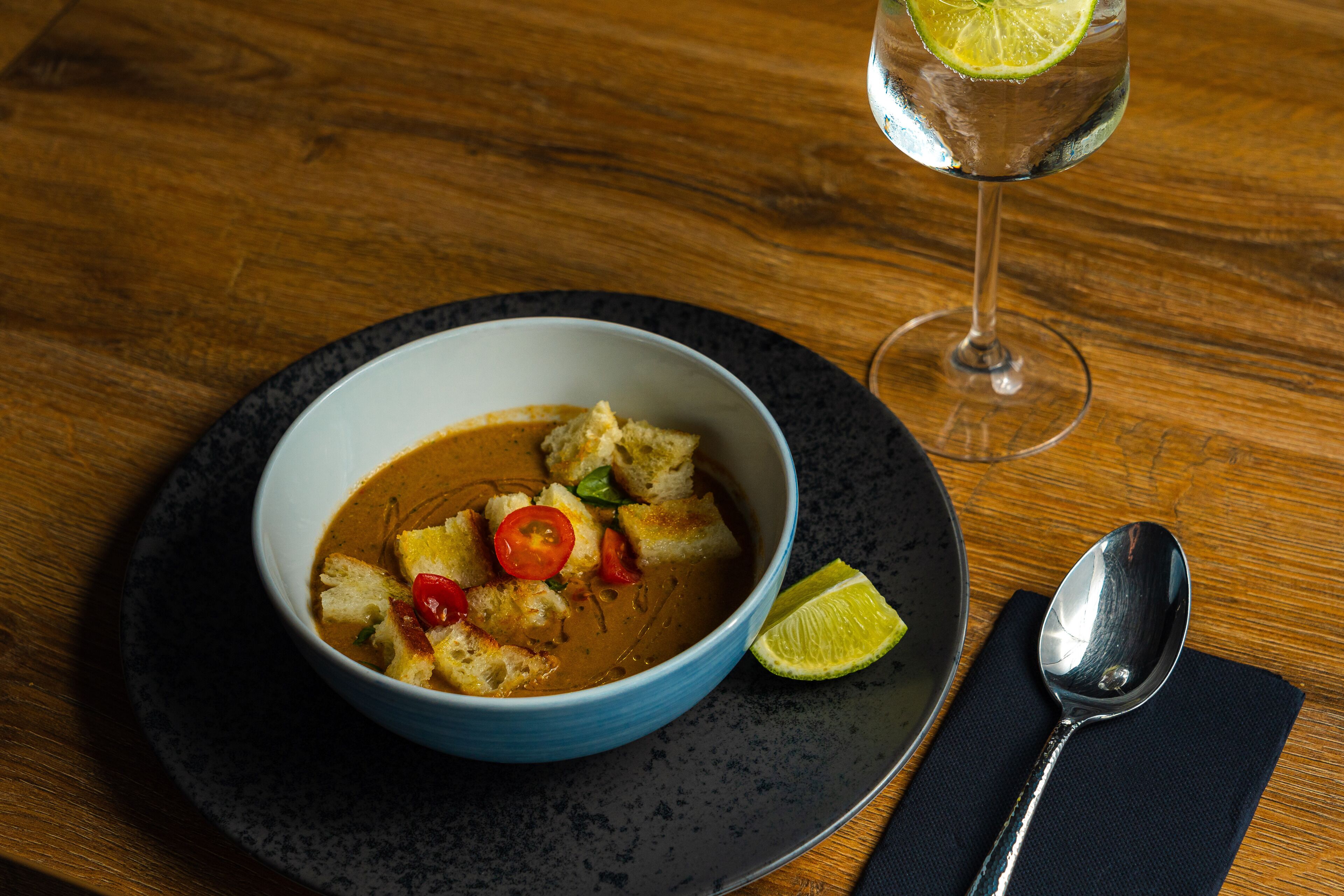 A bowl of soup with bread and lime on a plate, accompanied by a glass of water and a spoon on a napkin.