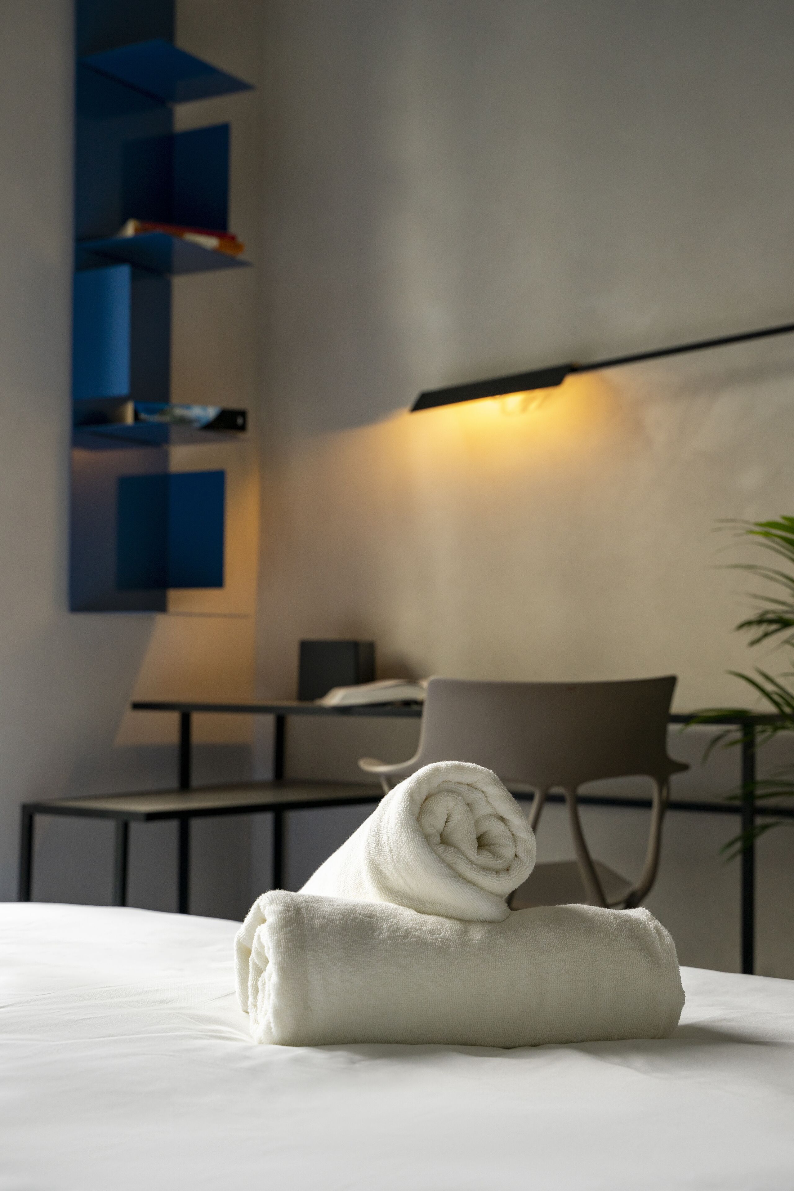 A neatly made bed with a rolled white towel, a modern chair, and a desk with a lamp in a contemporary room.