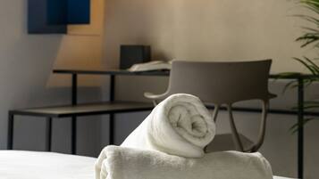 A neatly made bed with a rolled white towel, a modern chair, and a desk with a lamp in a contemporary room.