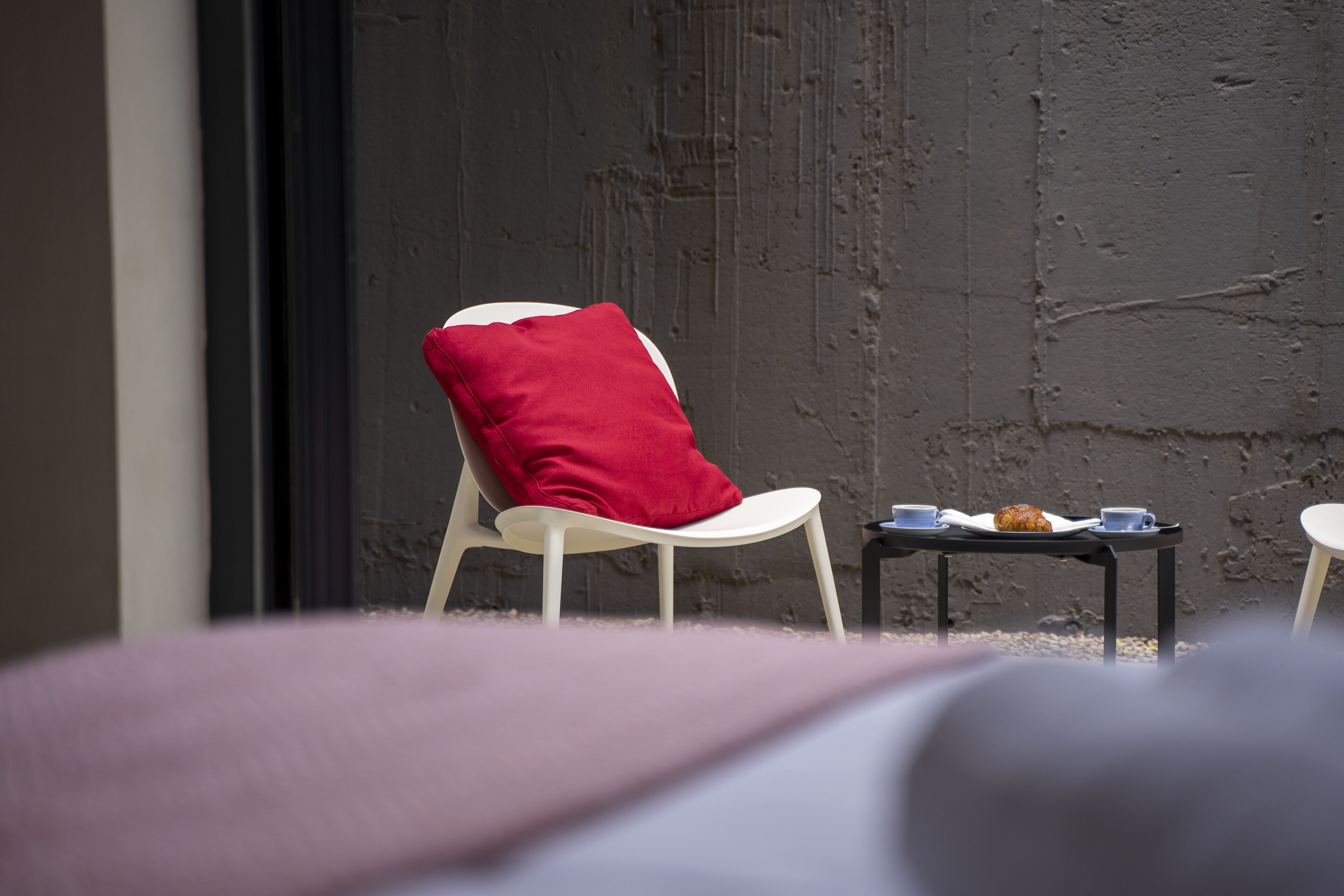 A white chair with a red pillow, a small table with a plate and cups, and a textured wall.