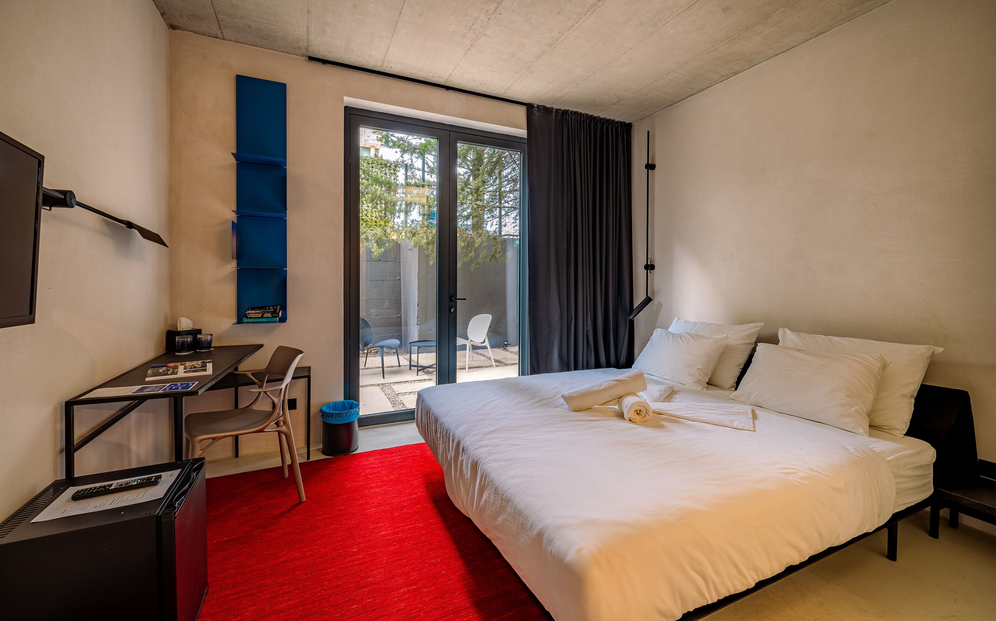 Hotelkamer met een bed, een bureau en een stoel. Er ligt een rood tapijt en er is een raam met gordijnen.