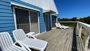 A deck with two white striped lounge chairs and a small table, a blue building with large windows, and a clear sky. - After Dune Delight, Indulge in your Ocean views. (Inverloch)