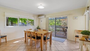 A dining area with a wooden table and chairs, a view of greenery outside, and a sliding glass door leading to an outdoor deck. - Surf Mist Retreat (Inverloch)