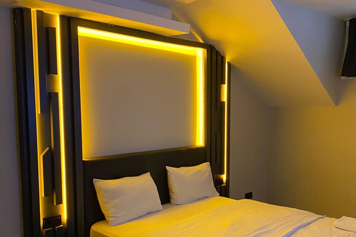 A modern bedroom with a large bed, a headboard with integrated lighting, and a wooden floor.
