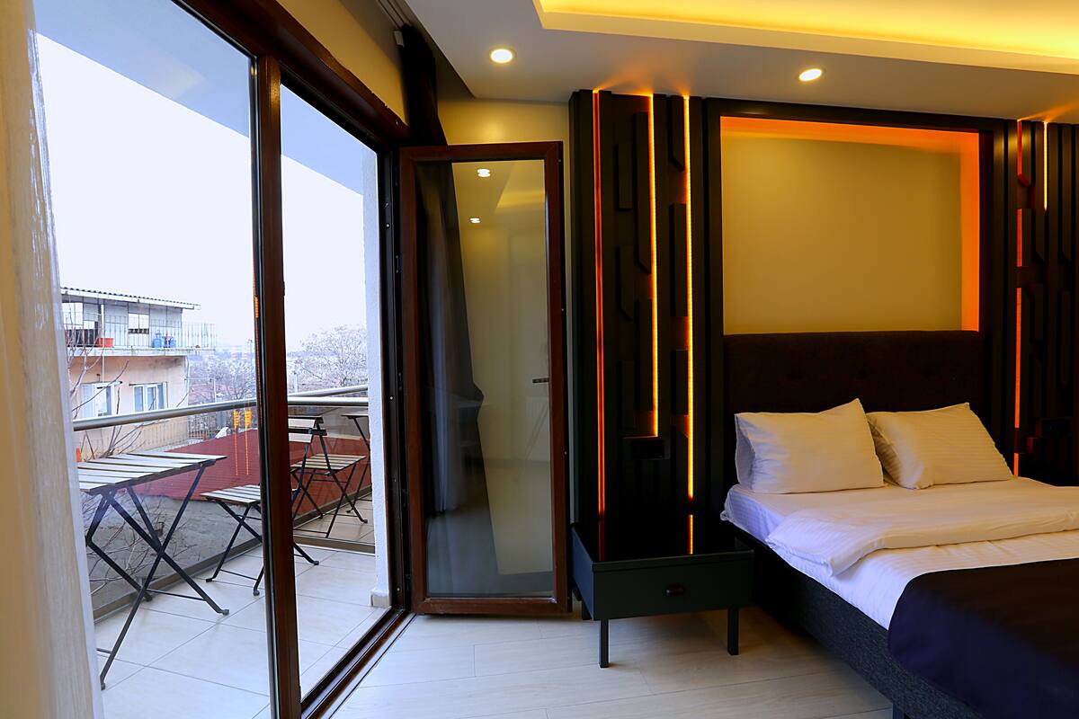 A modern hotel room with a large bed, a glass door leading to a balcony, and a view of the cityscape.