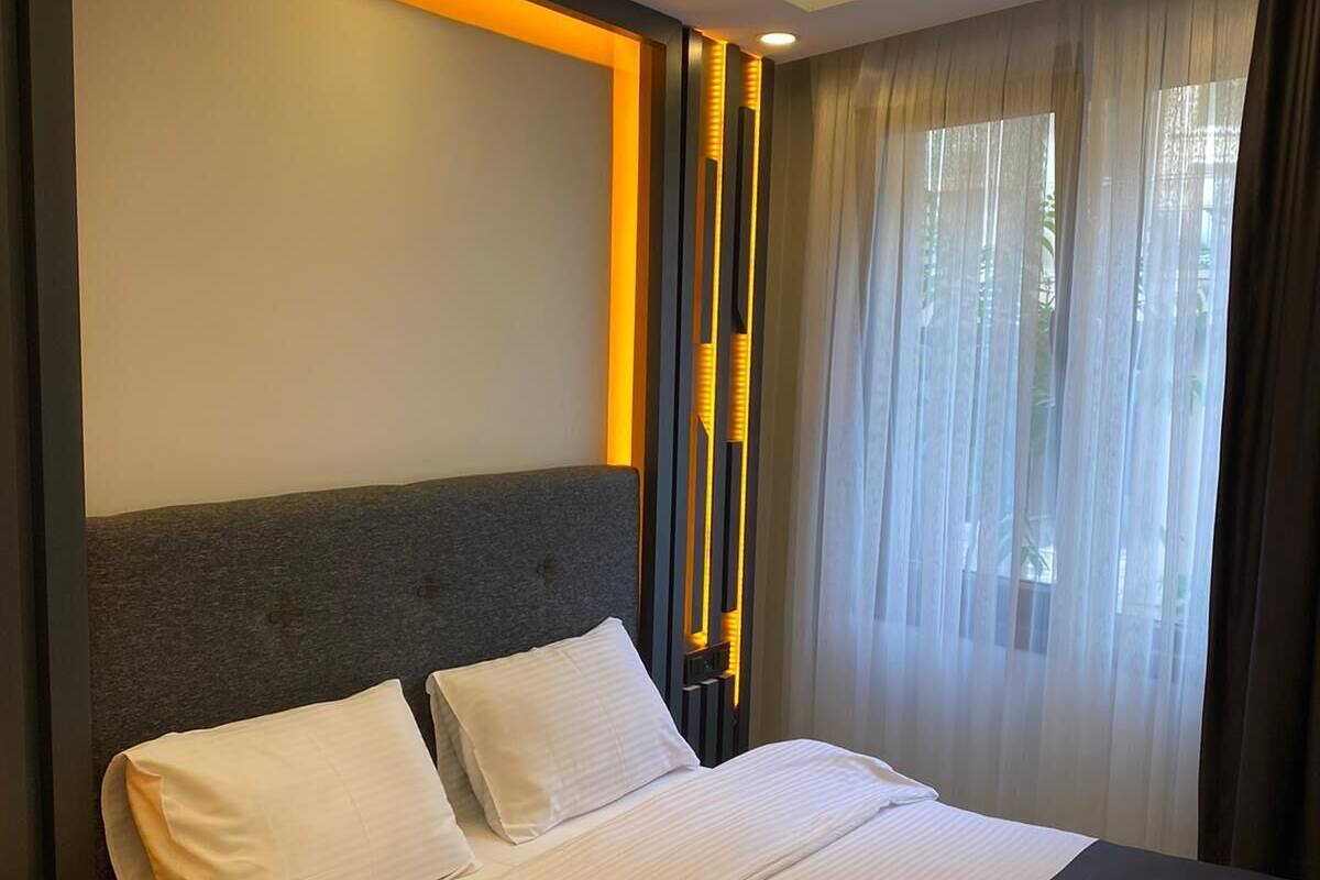 A modern hotel room with a large bed, a nightstand, and a window with sheer curtains.