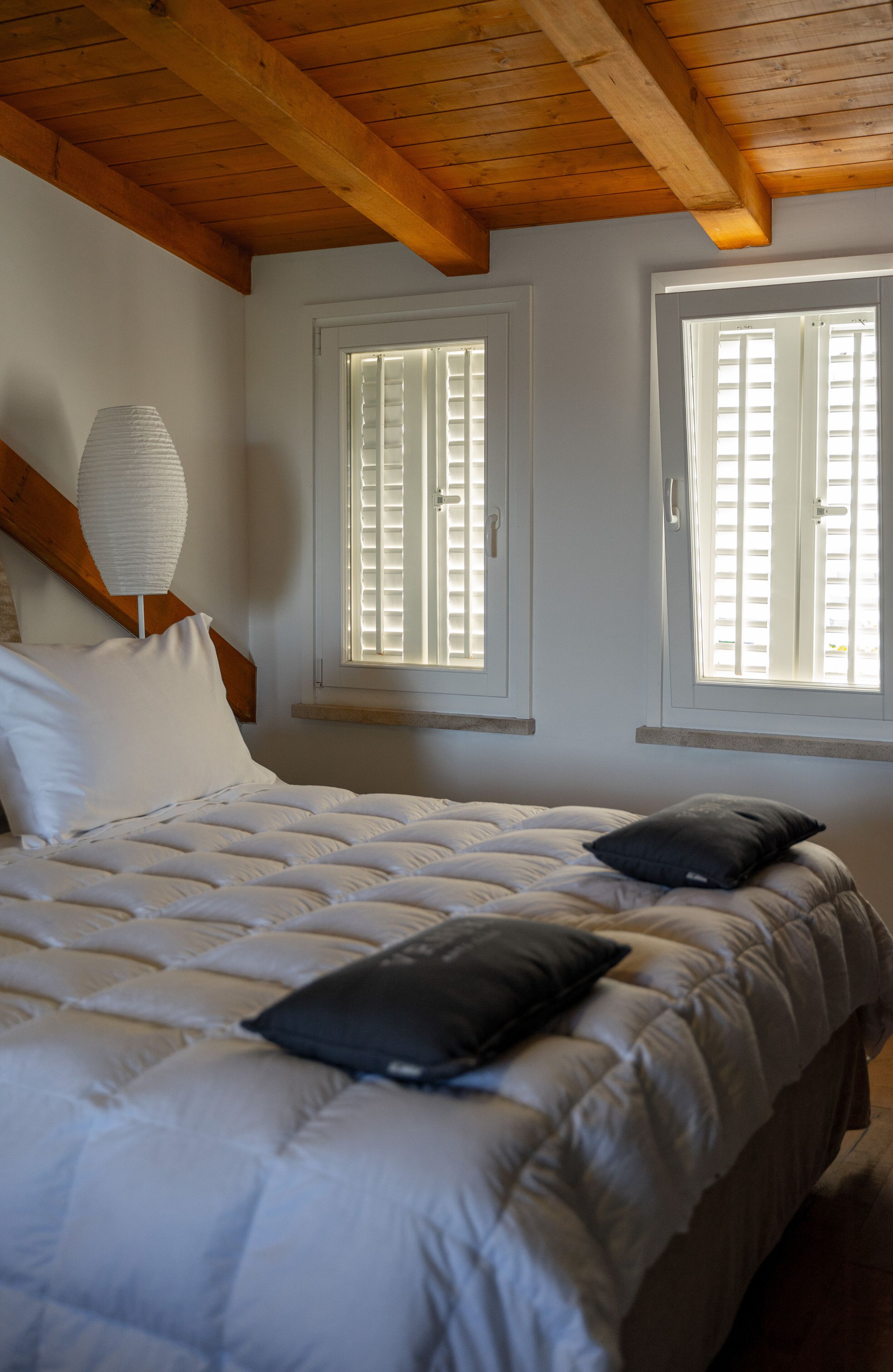 A bedroom with a bed, two windows with shutters, a wooden ceiling, and a bedside lamp.
