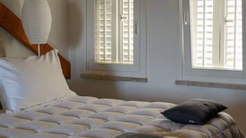 A bedroom with a bed, two windows with shutters, a wooden ceiling, and a bedside lamp.