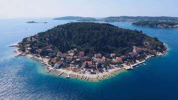 Vista aérea de un pueblo costero con una isla boscosa y densa, aguas azul turquesa y un pequeño puerto con barcos.