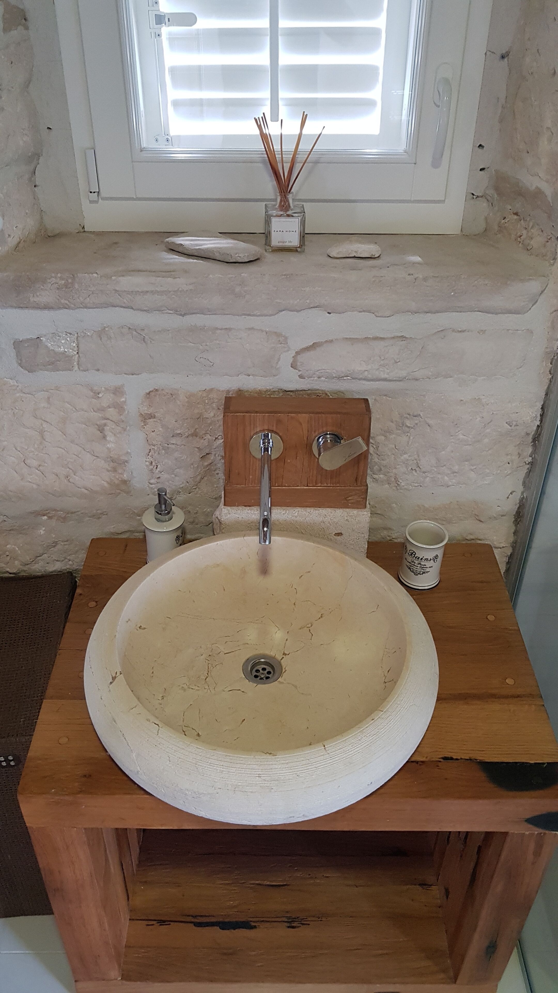 A bathroom with a round, beige sink on a wooden base, a wooden medicine cabinet above it, and a window with blinds in the background.