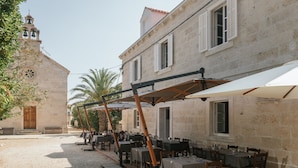 Un área de comedor al aire libre con mesas y sillas, una estructura de pérgola y un edificio con una torre de campanas al fondo.