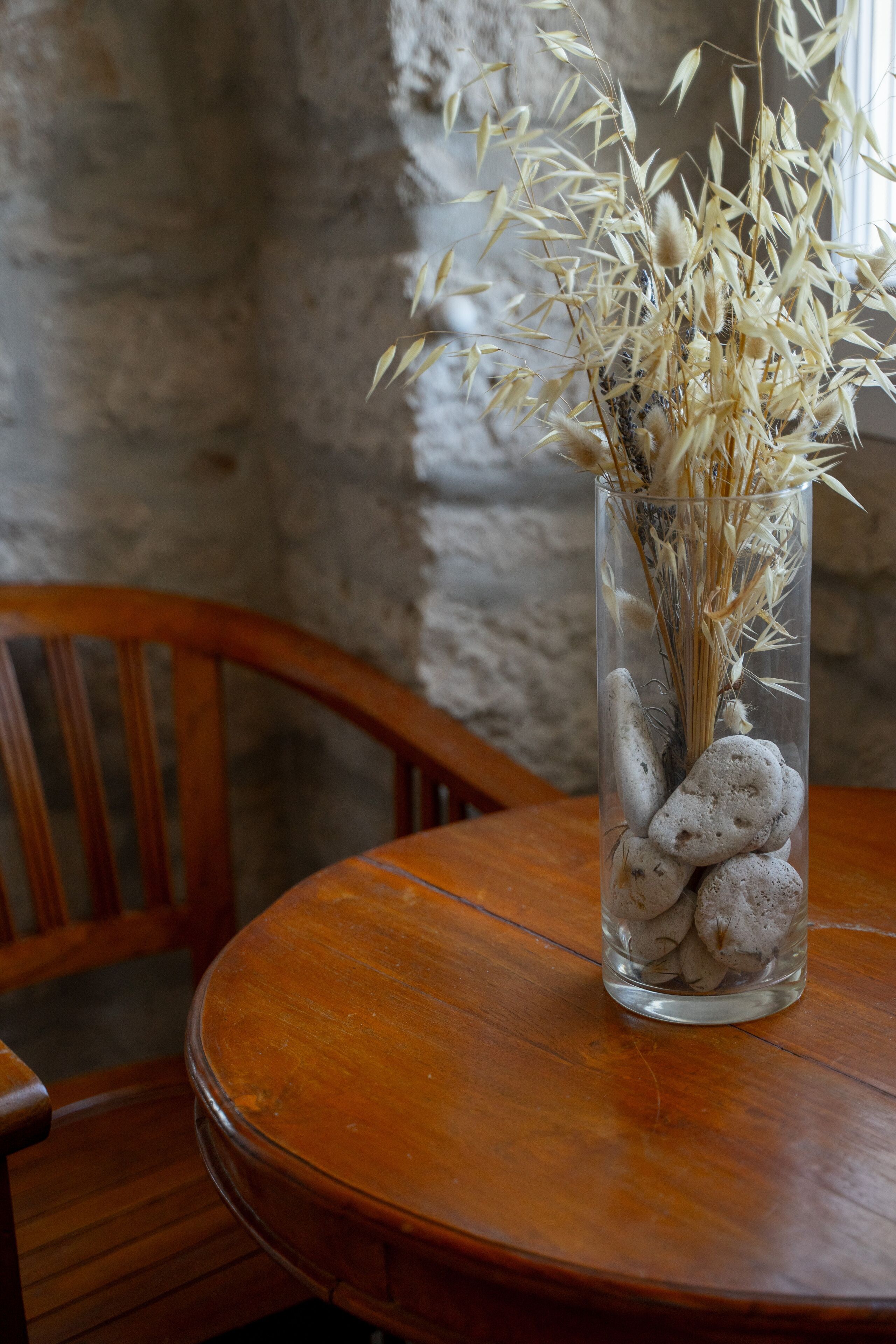 A clear glass vase with dried plants and smooth stones on a wooden table.