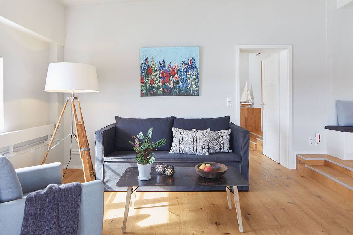 A modern living room with a dark blue sofa, a small coffee table with a plant and fruit bowl, a floor lamp, and a painting on the wall.