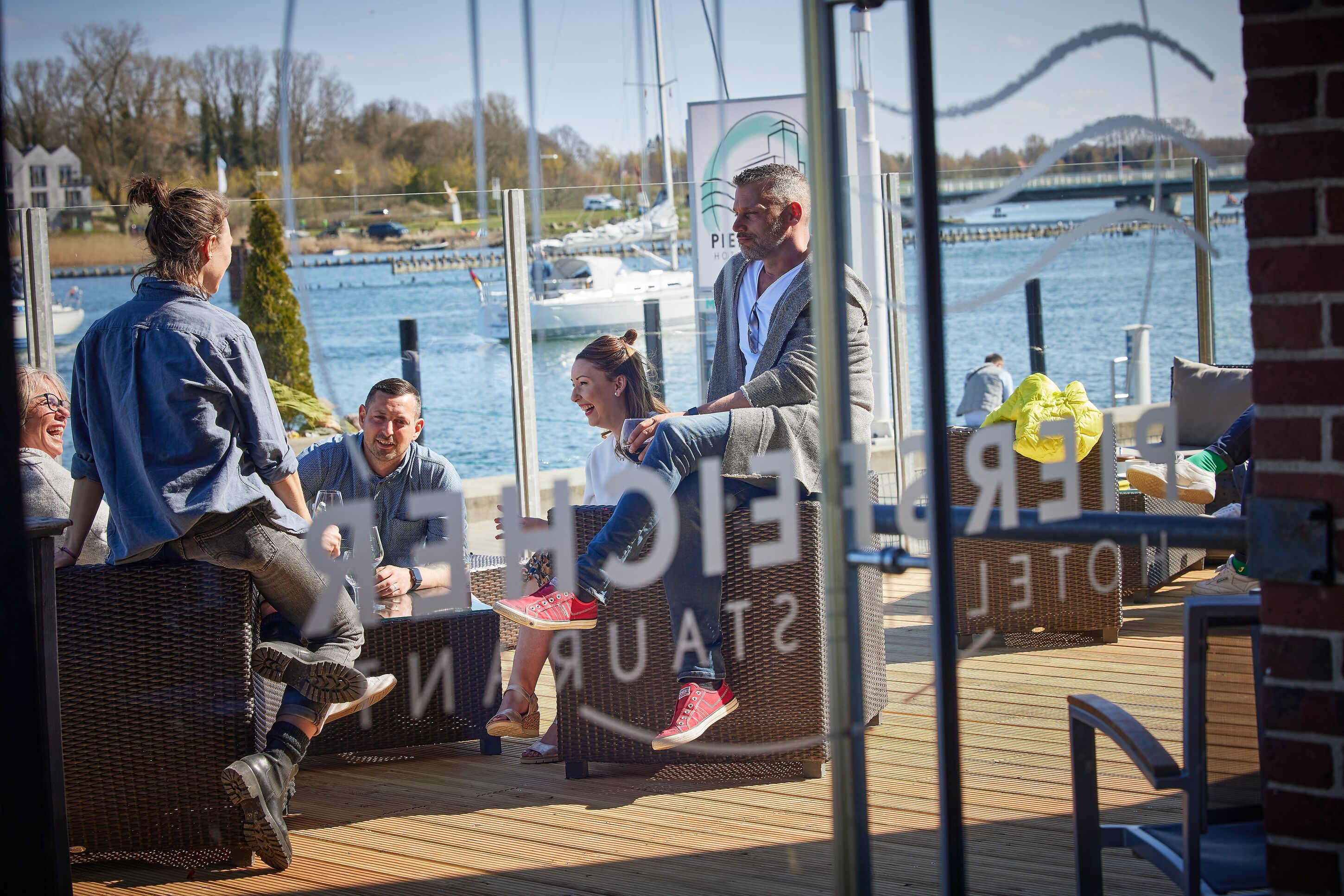 A group of people sitting on wicker furniture by a waterfront, overlooking a marina with boats and a bridge in the background.
