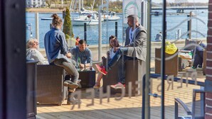 A group of people seated on wicker furniture on a wooden deck with a marina view in the background. - Pierspeicher Boardinghouse (Kappeln)