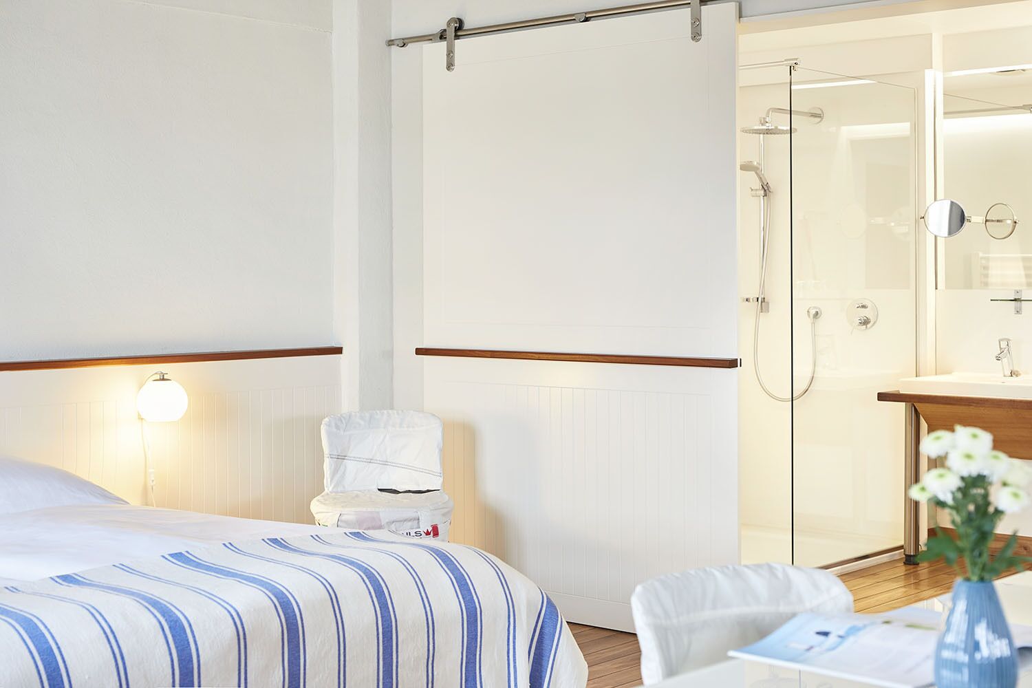A hotel room with a bed, a bedside table with a lamp, a shower area, and a bathroom with a sink and mirror.