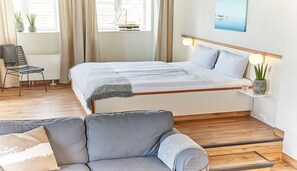 A bedroom with a bed, a chair, a sofa, a small table, and a window with curtains. - Pierspeicher Boardinghouse (Kappeln)