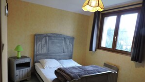 Ein Schlafzimmer mit einem hölzernen Bett, einem Nachttisch mit Lampe, einem Fenster mit Vorhängen und einer Heizung.
