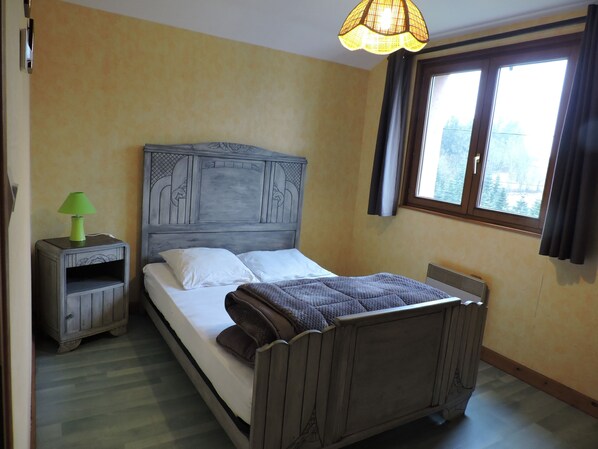 Ein Schlafzimmer mit einem hölzernen Bett, einem Nachttisch mit Lampe, einem Fenster mit Vorhängen und einer Heizung.