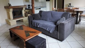 Ein Wohnzimmer mit einer Couch, einem Couchtisch und einem Kamin. Im Hintergrund ist eine Küche mit Schränken und einem Herd zu sehen.