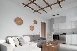 A modern living room with a grey sofa, wooden beams on the ceiling, a kitchen with white cabinets, and a wooden coffee table.