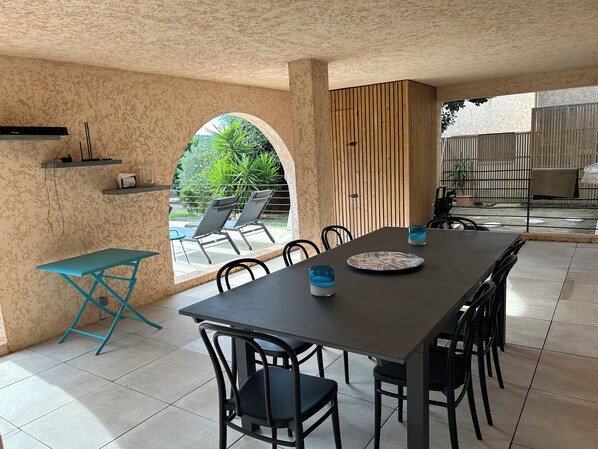 A dining area with a long table, chairs, a blue folding table, and a view of a garden through an arched window.