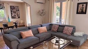 Un salon moderne avec un canapé d’angle gris, une table basse en verre et un coin repas comprenant une table et des chaises.