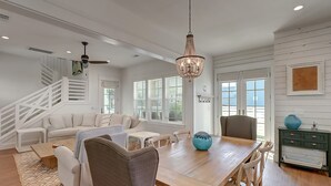 A spacious living area with a wooden dining table, comfortable seating, a staircase, and large windows allowing natural light. - Serene Living in Cinnamon Shore, w/ Access to Multi Pools & More Amenities (Port Aransas)
