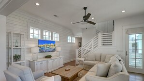 A spacious living room with a white wooden wall, a large flat-screen TV, a ceiling fan, and a staircase. - Serene Living in Cinnamon Shore, w/ Access to Multi Pools & More Amenities (Port Aransas)