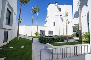 Un bâtiment moderne aux murs extérieurs blancs, agrémenté de palmiers et doté d’une pelouse bien entretenue.